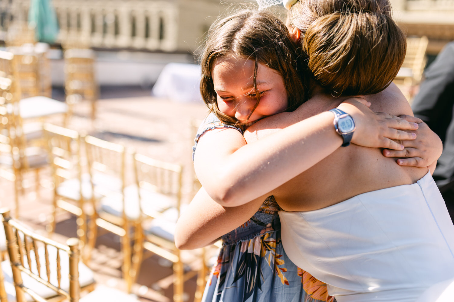 Two women sharing a heartfelt hug outdoors, surrounded by elegant golden chairs in a festive atmosphere.