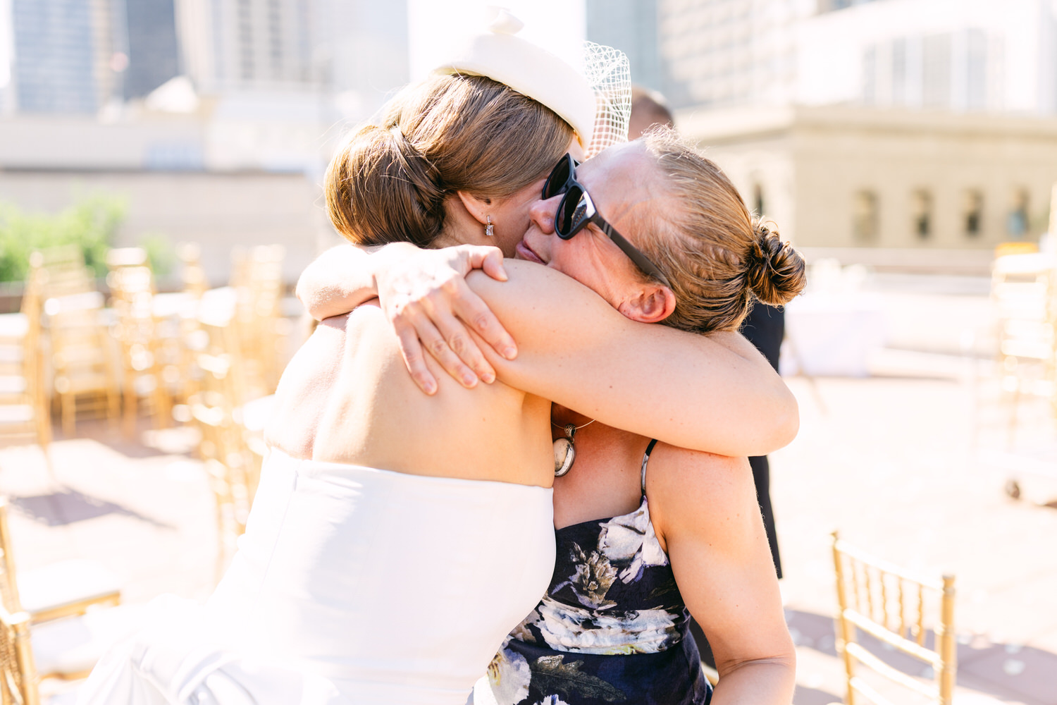 A joyful moment capturing two women embracing during a wedding celebration on a sunny rooftop, with elegant gold chairs in the background.