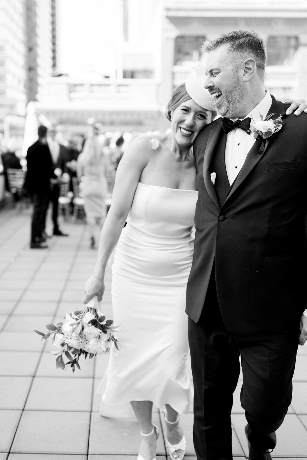 A joyful couple poses close together, the woman in a sleek white dress and a headpiece, and the man in a classic tuxedo, both smiling amidst a lively outdoor wedding reception.