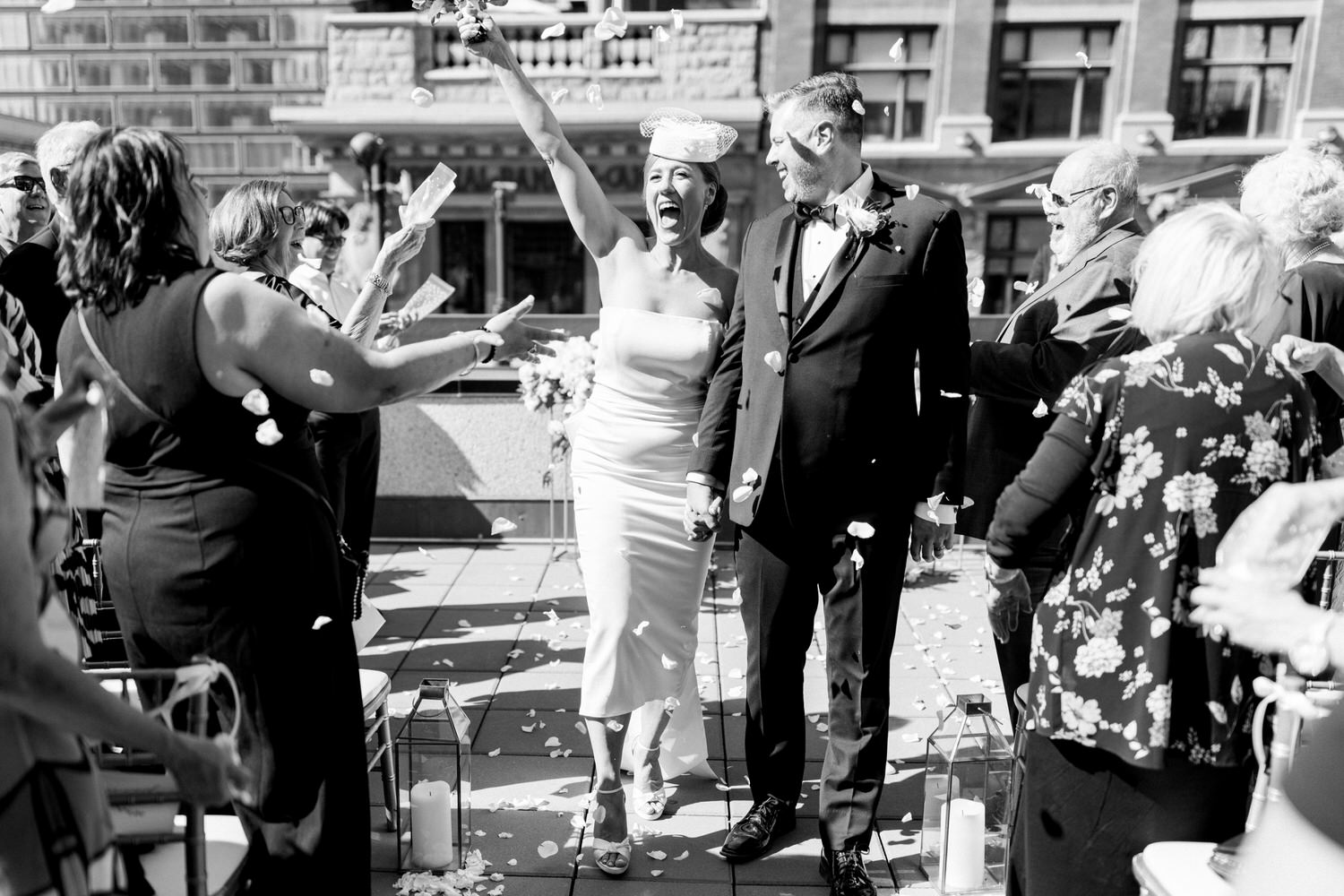 A happy couple walks hand in hand as guests shower them with flower petals at their outdoor wedding ceremony.