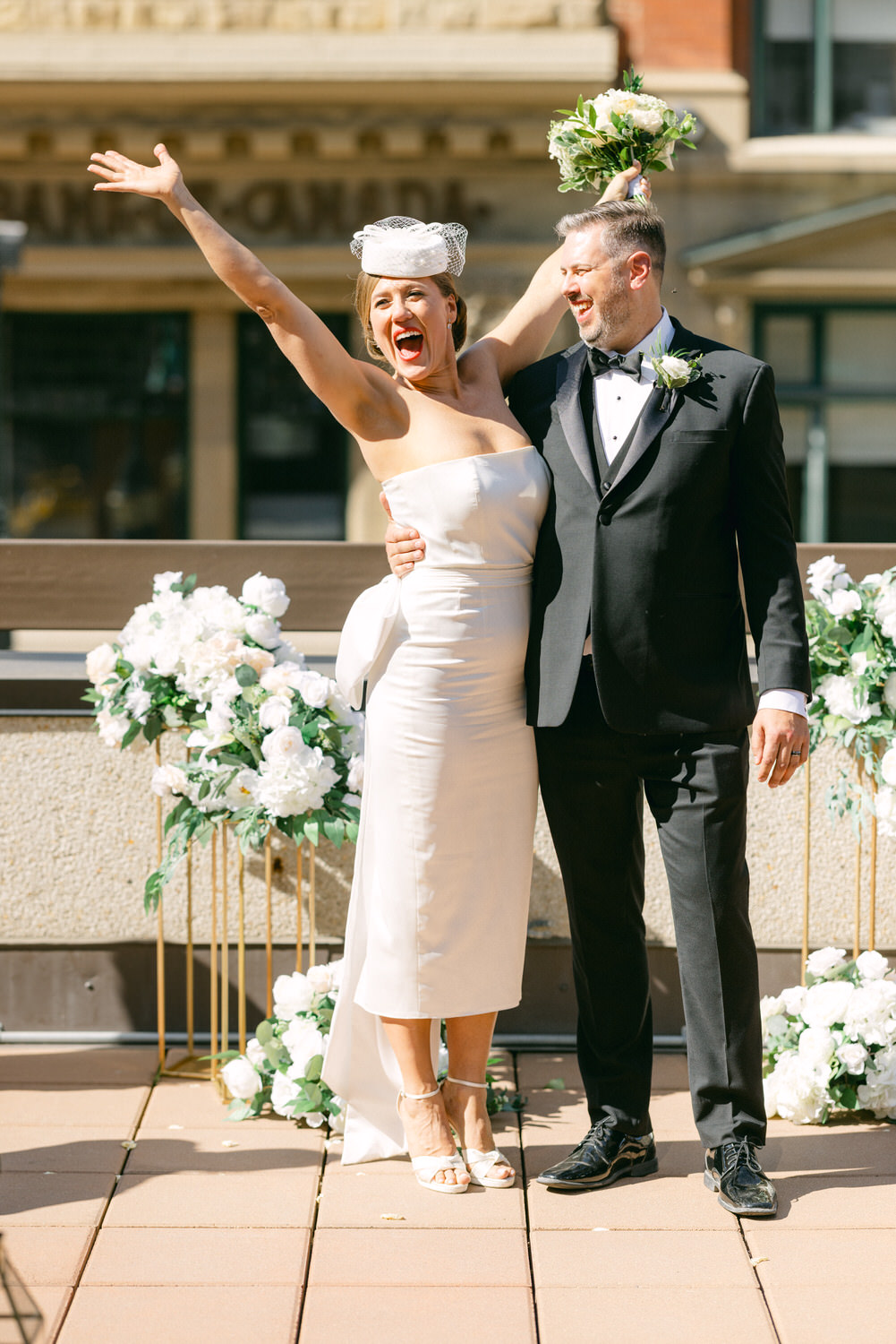 A happy couple celebrates their wedding, with the bride exuberantly raising her arms and holding a bouquet, while the groom stands beside her smiling, surrounded by floral decorations.