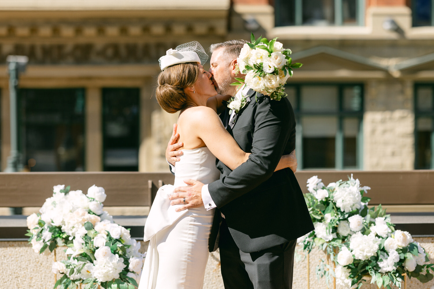 A couple embracing and sharing a kiss during their wedding ceremony, surrounded by floral arrangements in a charming outdoor venue.