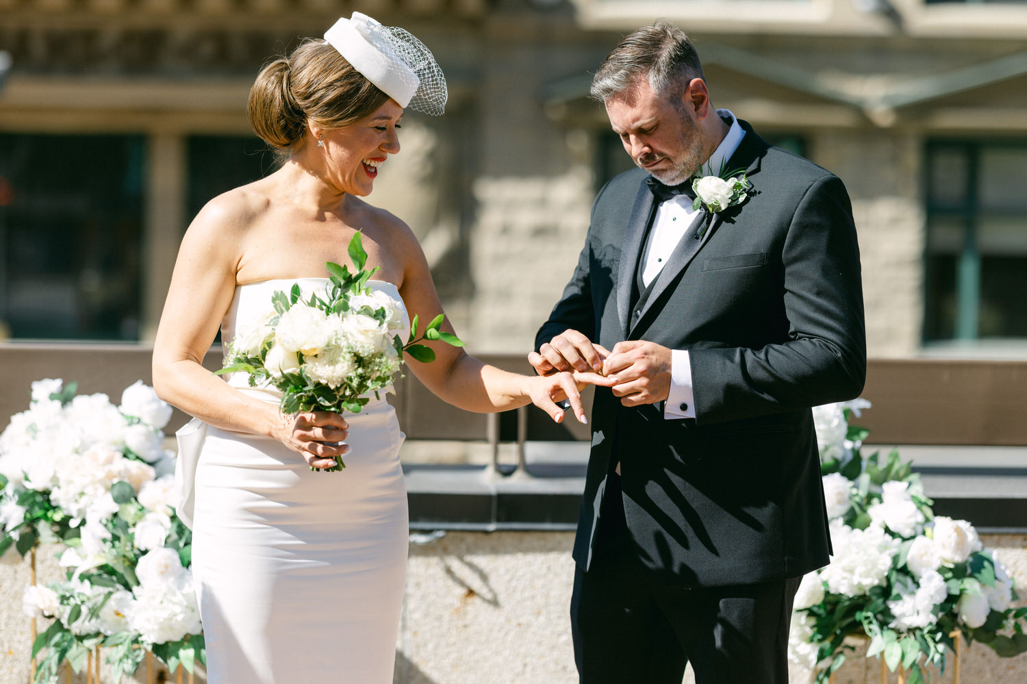 Bride smiling as groom places a ring on her finger, surrounded by floral decorations.