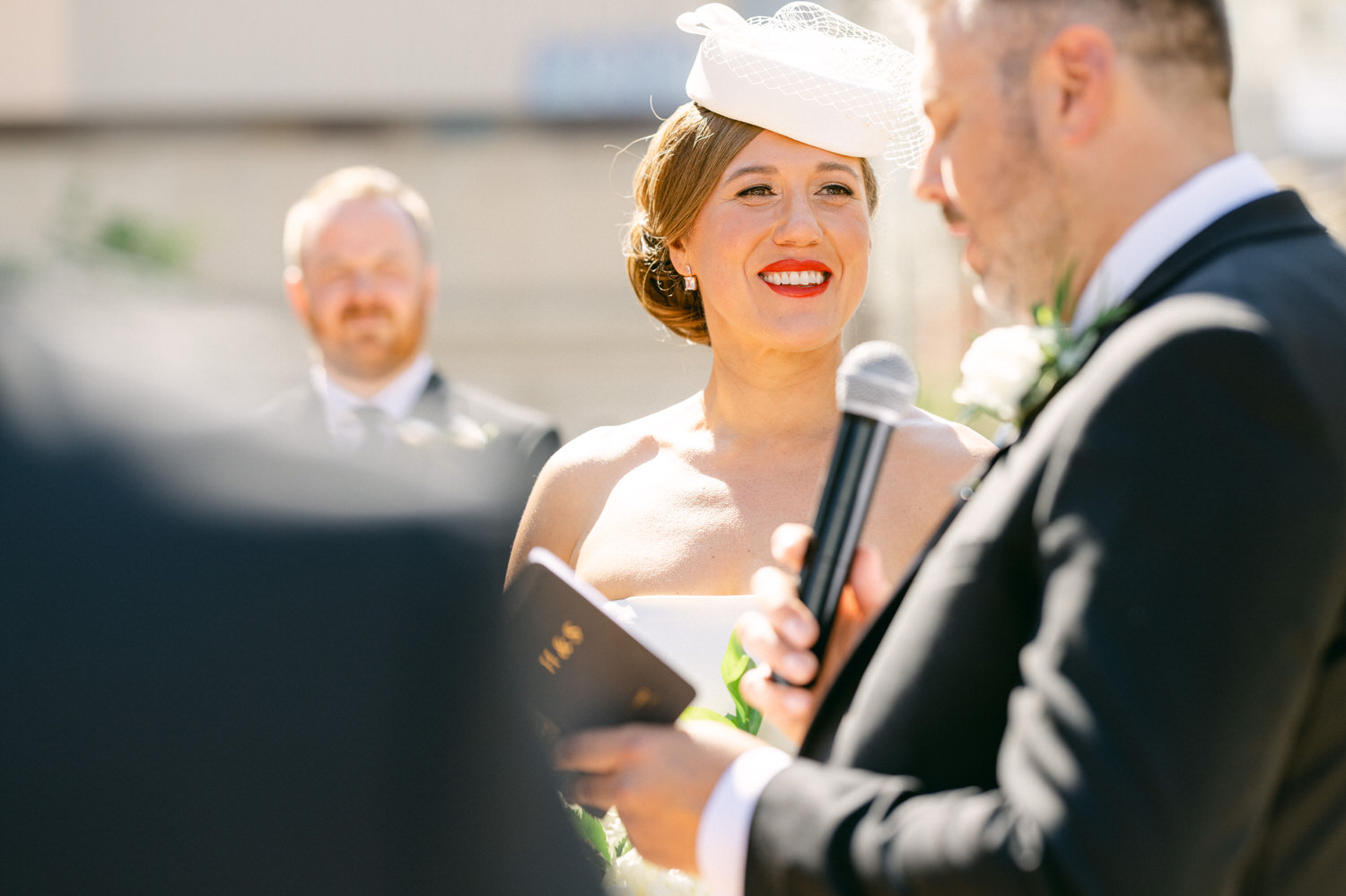 A radiant bride smiles during a wedding ceremony as the officiant speaks, captured in bright sunlight.