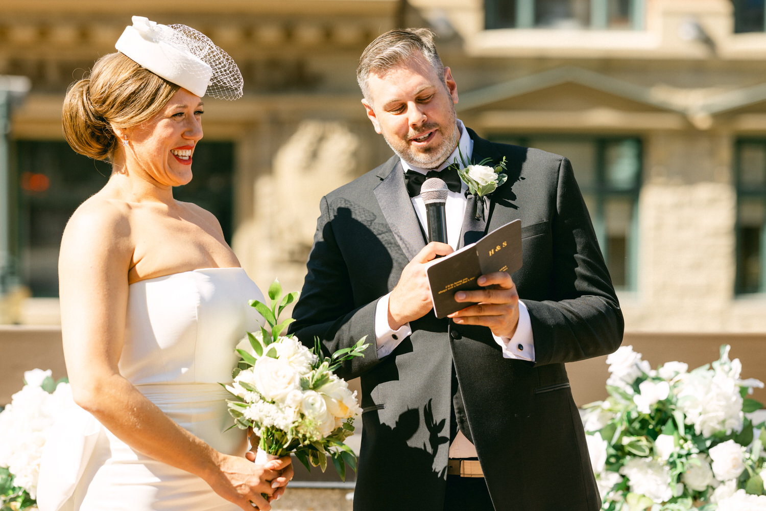 A bride in a strapless gown smiles as a suited officiant reads from a booklet during their wedding ceremony, surrounded by floral arrangements in a sunny outdoor setting.