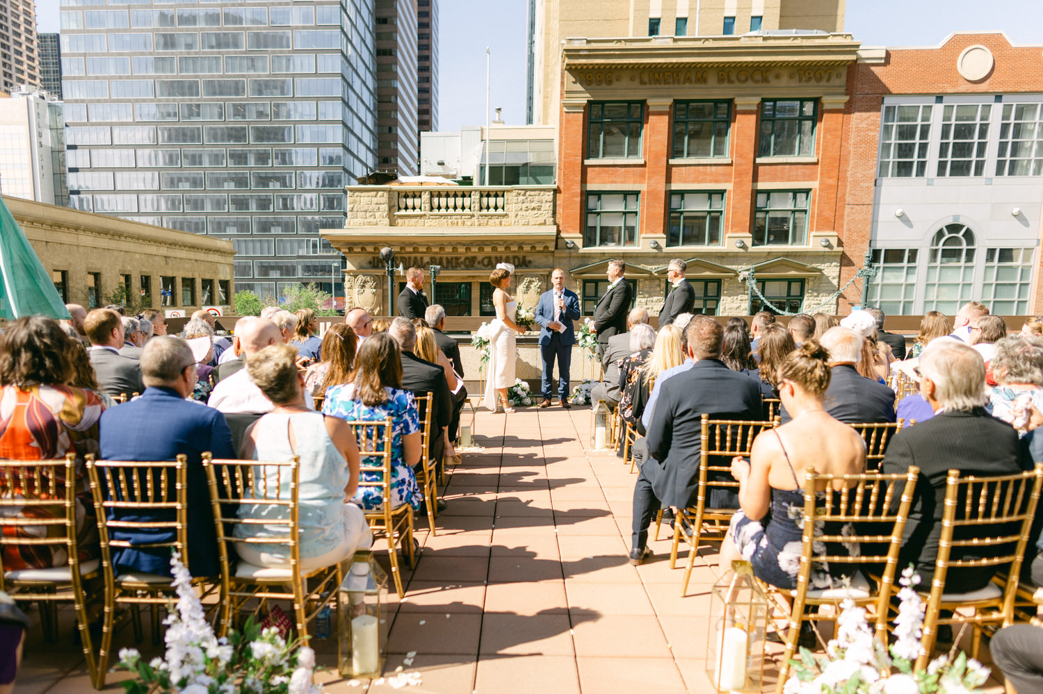 A couple exchanging vows during a beautiful outdoor wedding ceremony on a rooftop, surrounded by guests seated on elegant chairs with city buildings in the background.