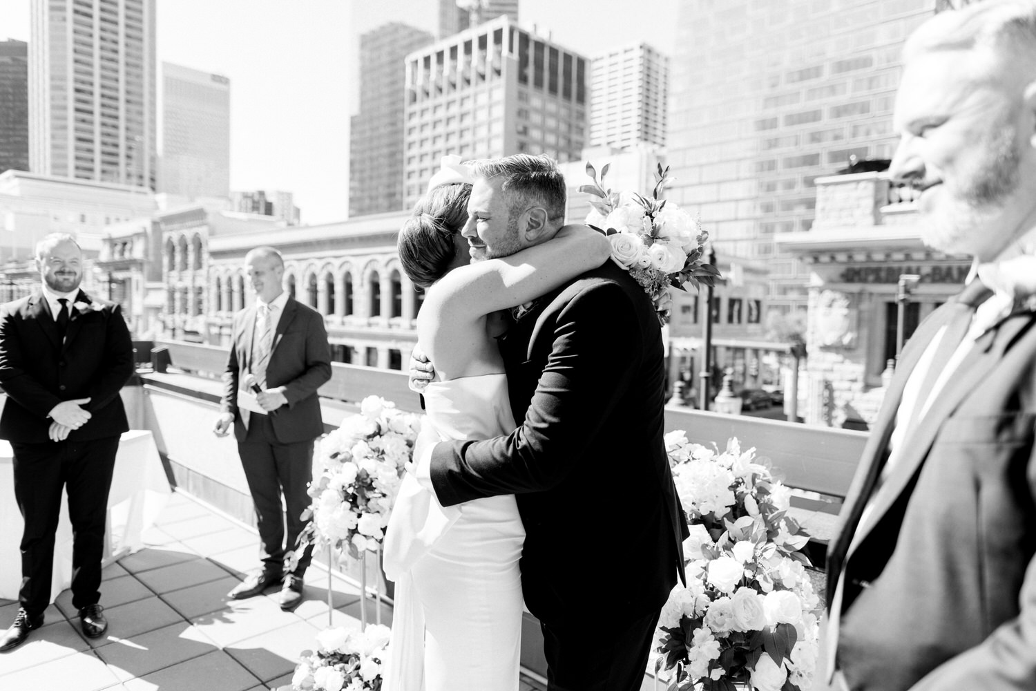 A bride and groom share a heartfelt hug during their wedding ceremony on a sunny city rooftop, surrounded by floral decorations and guests in the background.