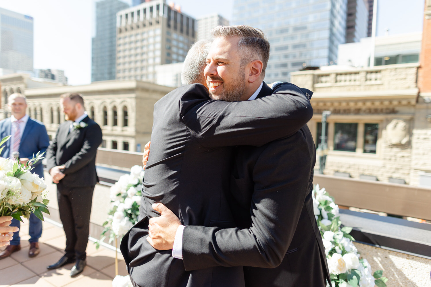 Two men embrace joyfully during a wedding celebration on a sunny rooftop, surrounded by floral arrangements and city buildings.