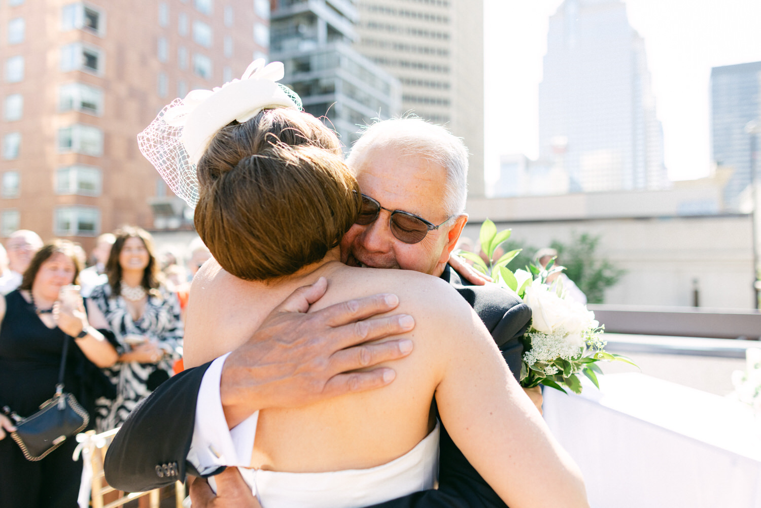 A bride in a stylish white gown shares a heartfelt hug with her father, surrounded by guests in a sunny outdoor setting.