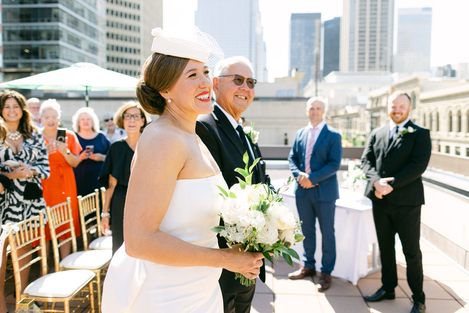 A bride smiles joyfully while holding a bouquet, accompanied by her father, at an outdoor wedding ceremony with guests and a city skyline in the background.