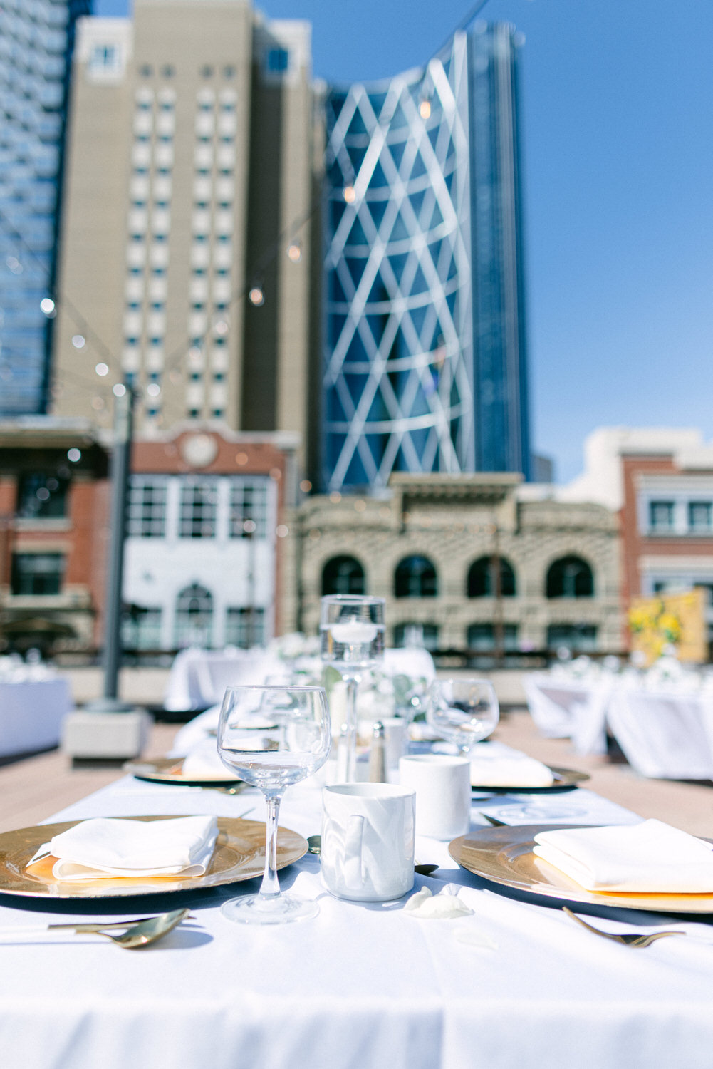 A beautifully arranged outdoor dining table featuring white tablecloths, gold plates, and elegant glassware, set against a backdrop of modern and historic buildings under a clear blue sky.