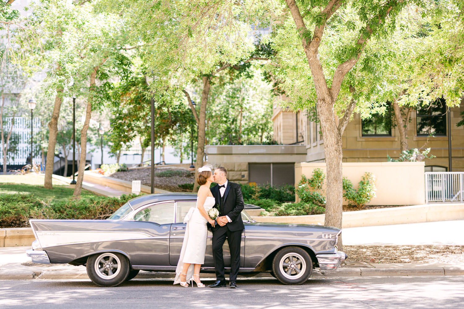 A bride and groom share a kiss beside a classic gray car in a lush outdoor setting, capturing a timeless wedding memory.