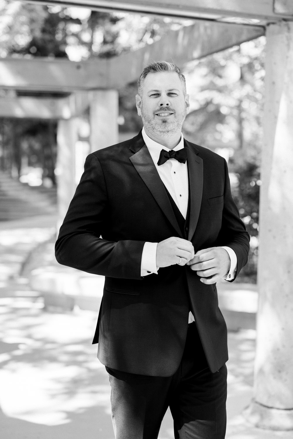 A groom in a tuxedo adjusting his bow tie, smiling confidently in an outdoor setting with soft lighting.