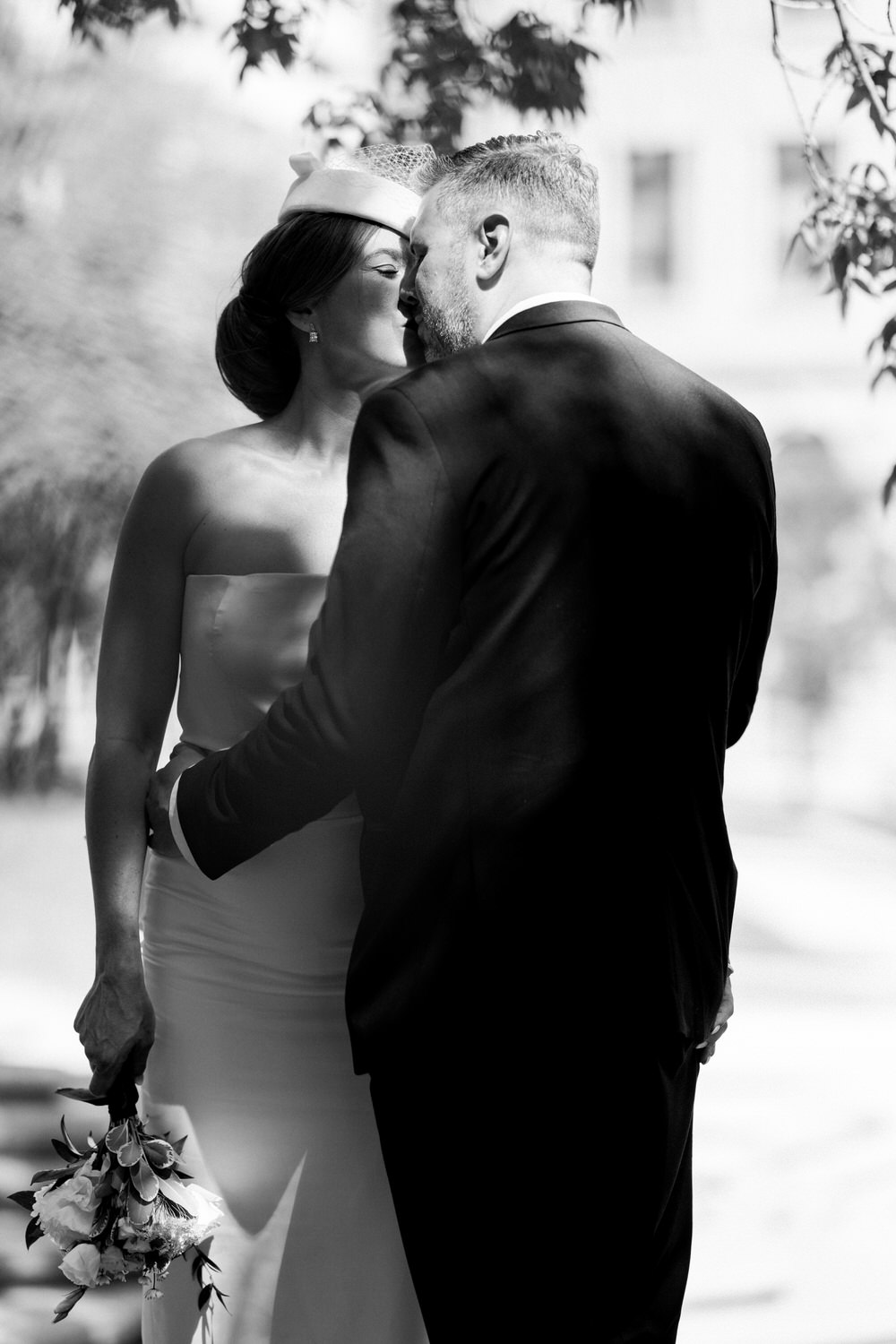 A bride and groom share a tender kiss in a black and white setting, showcasing intimate moments on their special day while holding a bouquet.