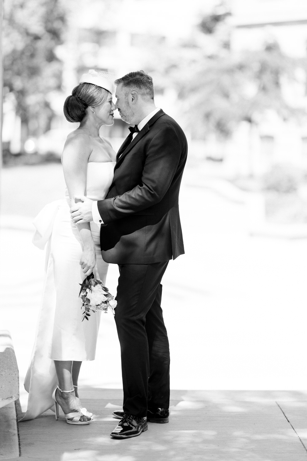 A bride and groom share a romantic moment, gazing into each other's eyes, with the bride holding a small bouquet and dressed in a sleek white gown, while the groom is in a classic black suit.