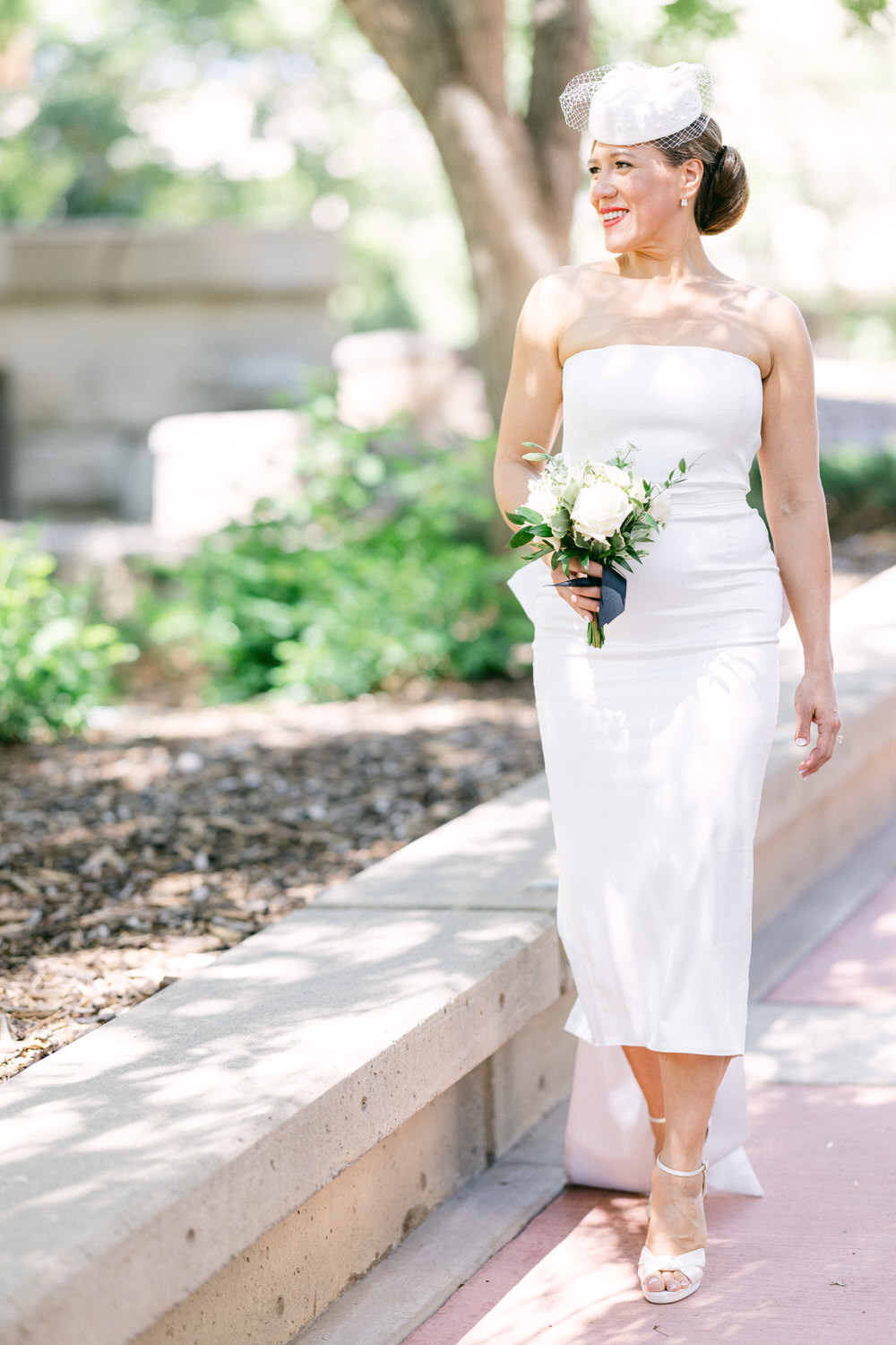 A smiling bride in a strapless white dress walking through a lush green park while holding a bouquet of white roses.