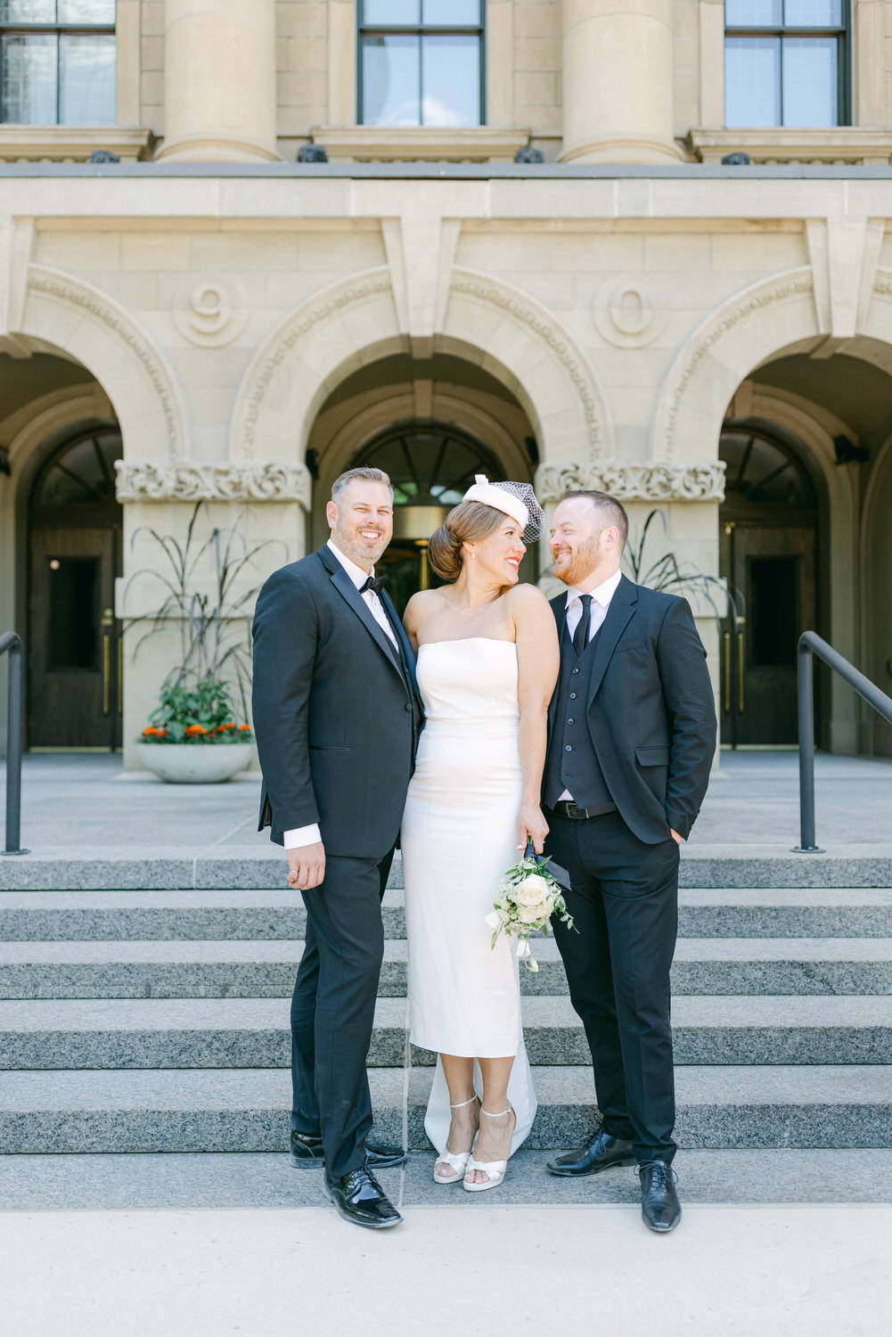 A bride in a strapless white dress poses joyfully with two suited friends on a staircase outside a historic building.