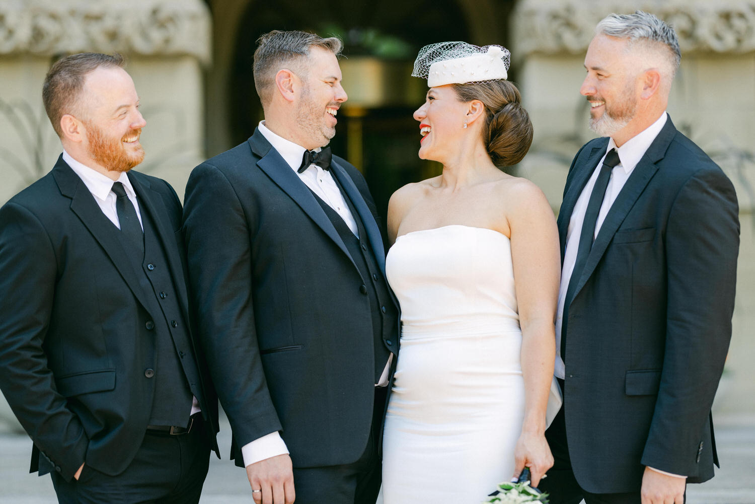 A joyful group of four, including a bride in a white dress and three men in formal suits, sharing smiles and laughter during a wedding celebration.