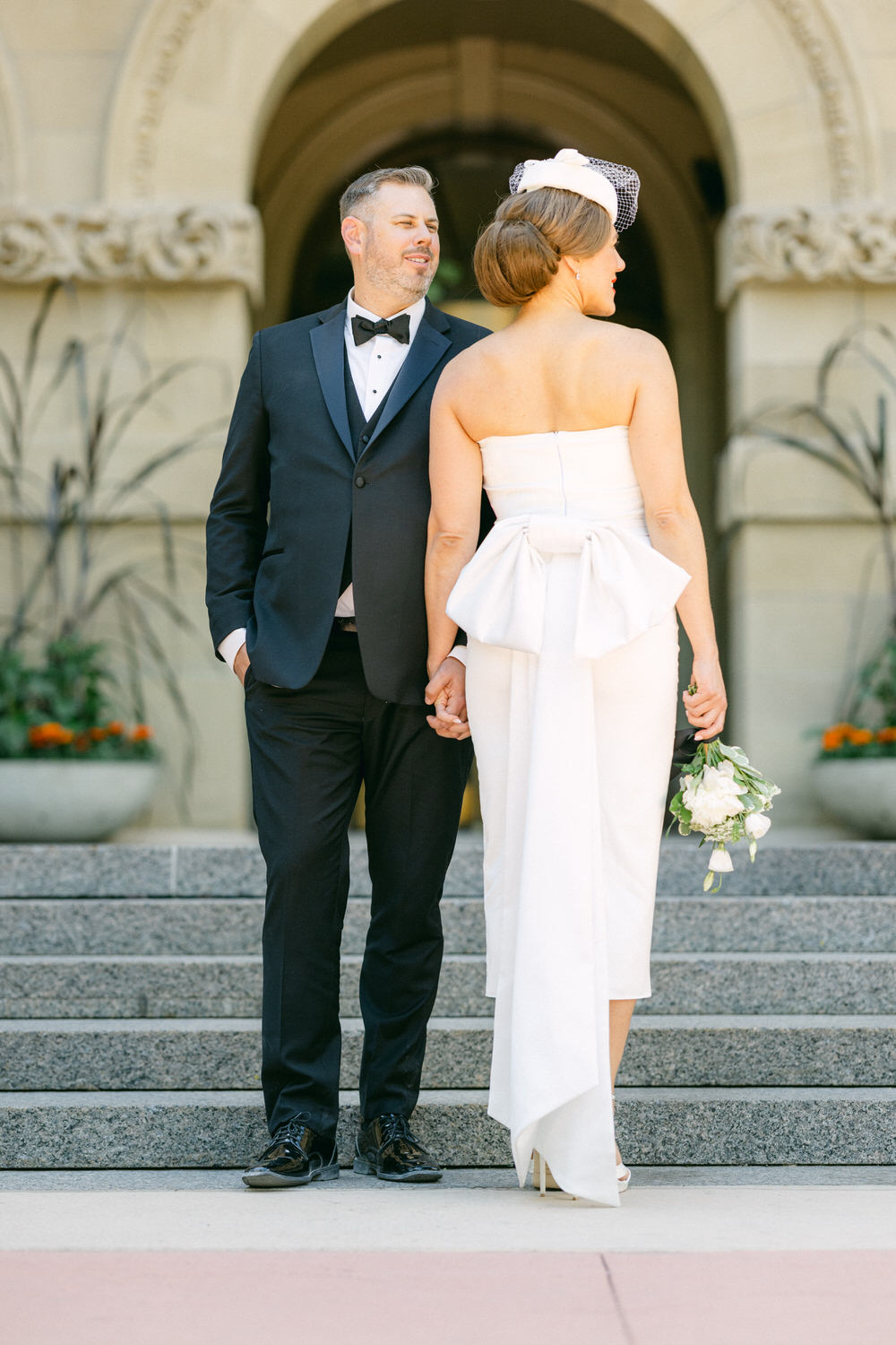 A couple in elegant wedding attire stands on steps, holding hands and gazing at each other, surrounded by architectural beauty.