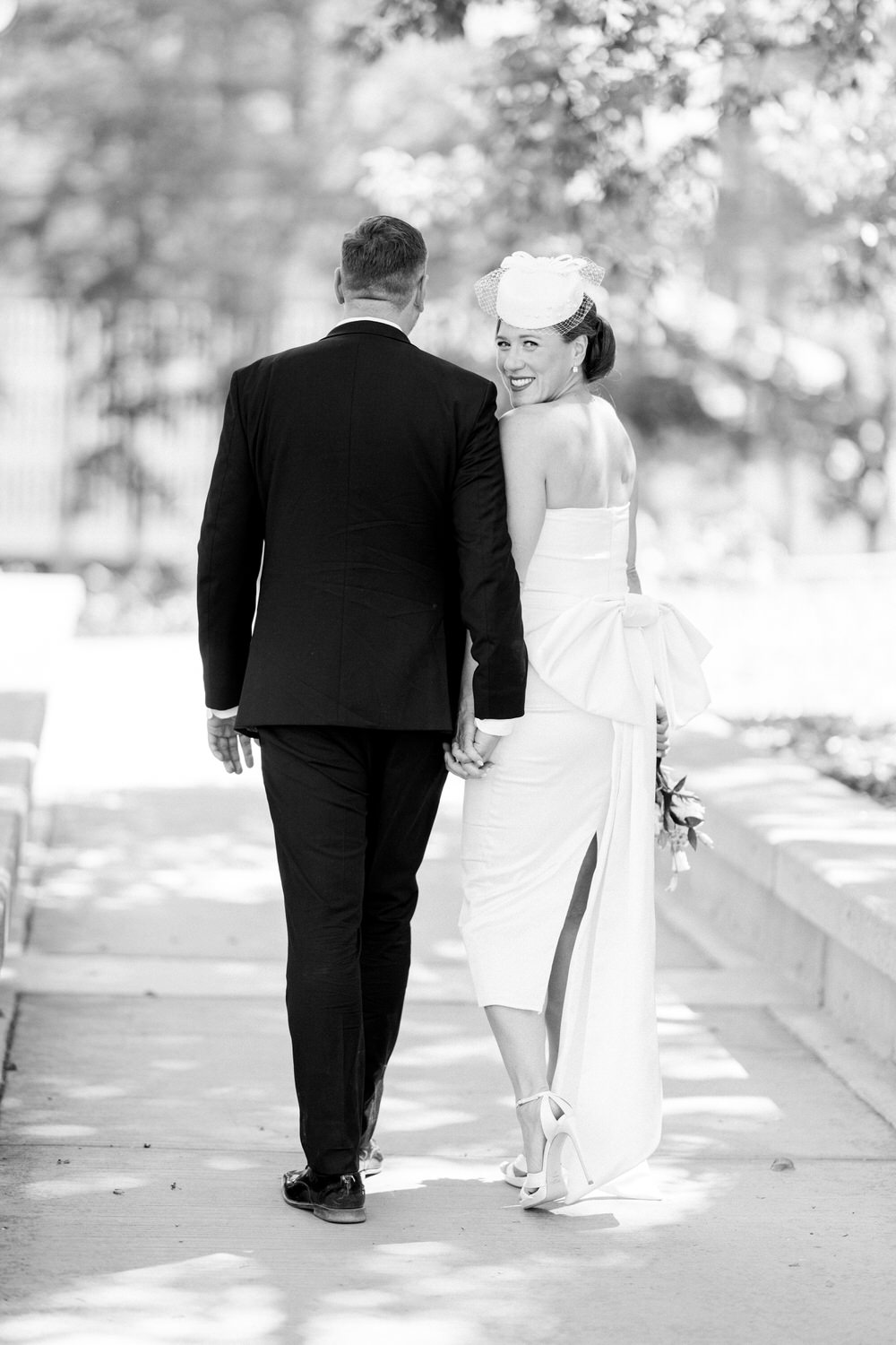 A couple walking hand in hand, with the bride glancing back and smiling, showcasing her elegant dress and headpiece in a picturesque outdoor setting.