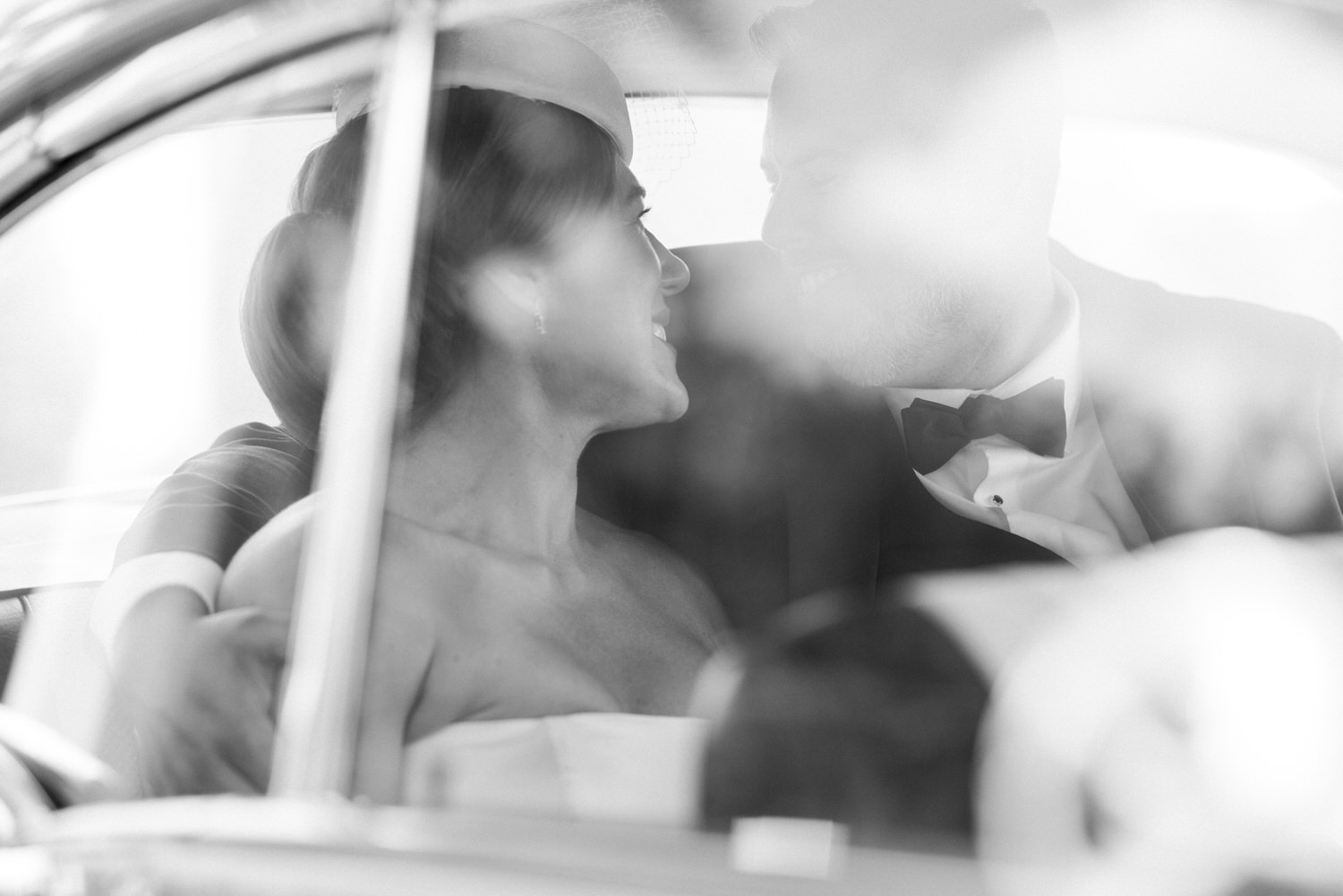 A bride and groom share a joyful gaze inside a classic car, captured in a soft black and white photograph, highlighting their love and connection.