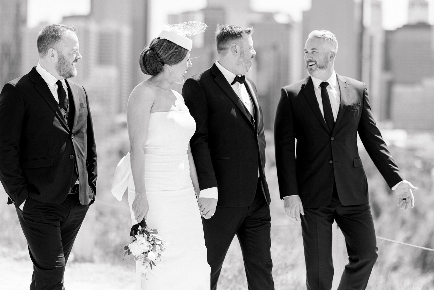 A stylish bride in a strapless gown and veil walks hand-in-hand with her groom and two groomsmen, showcasing joy and elegance against a city backdrop.