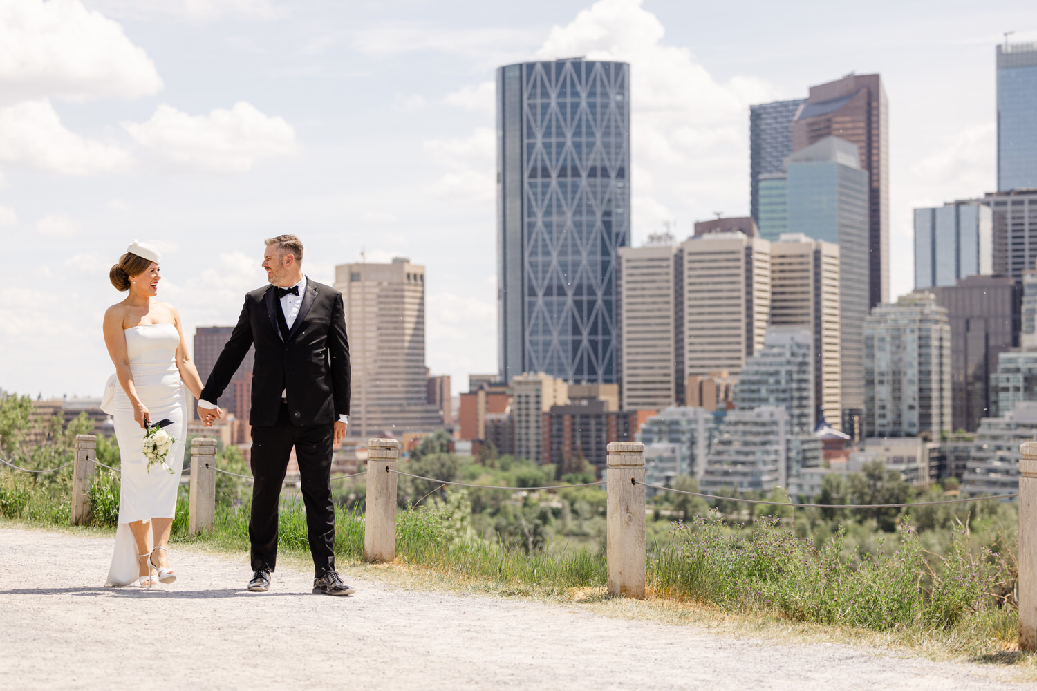 A bride and groom holding hands while walking along a scenic path with a city skyline in the background.