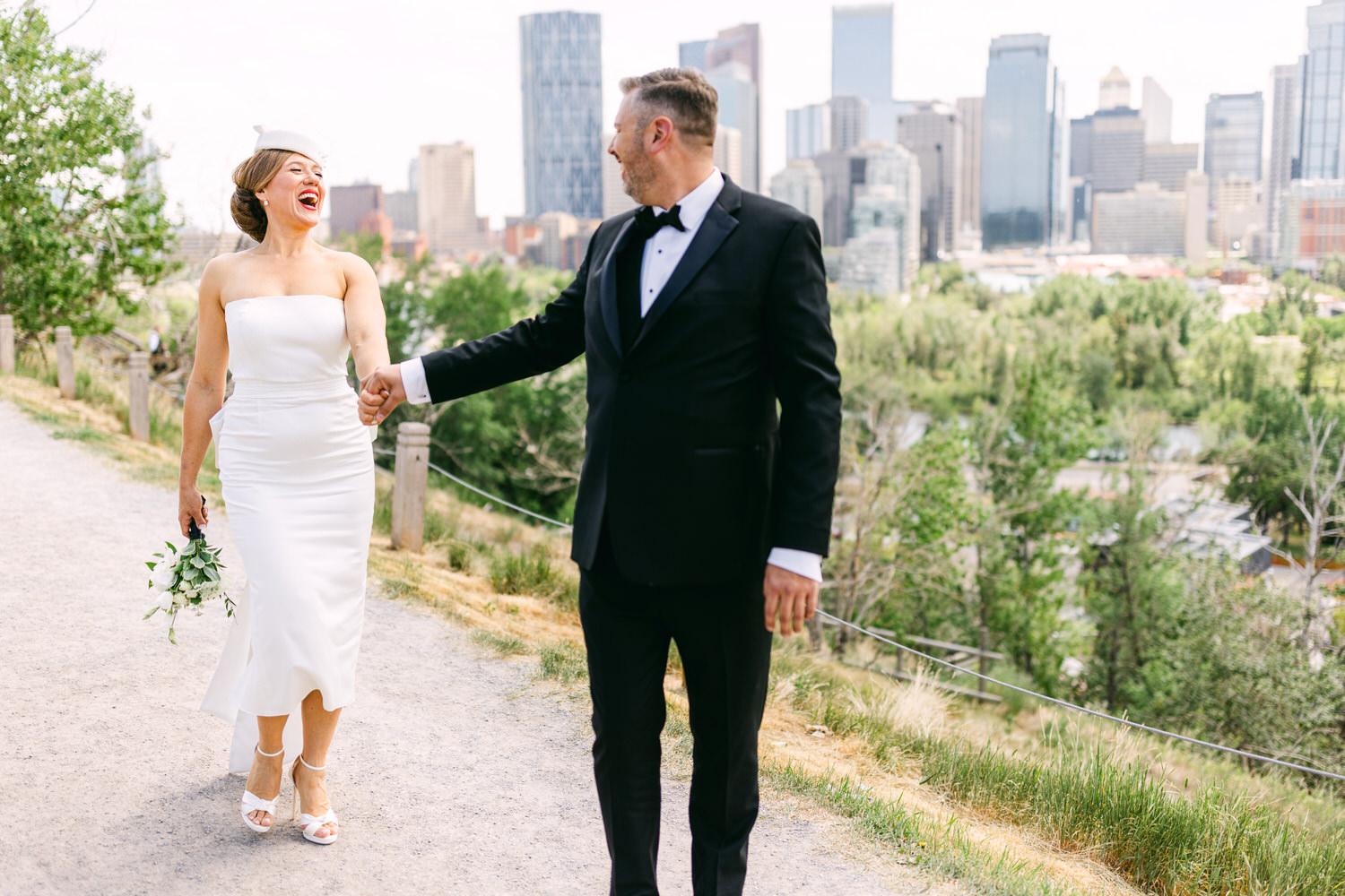 A happy bride and groom stroll hand in hand, with the bride in a sleek white dress and the groom in a black tuxedo, enjoying a scenic backdrop of a city skyline and greenery.