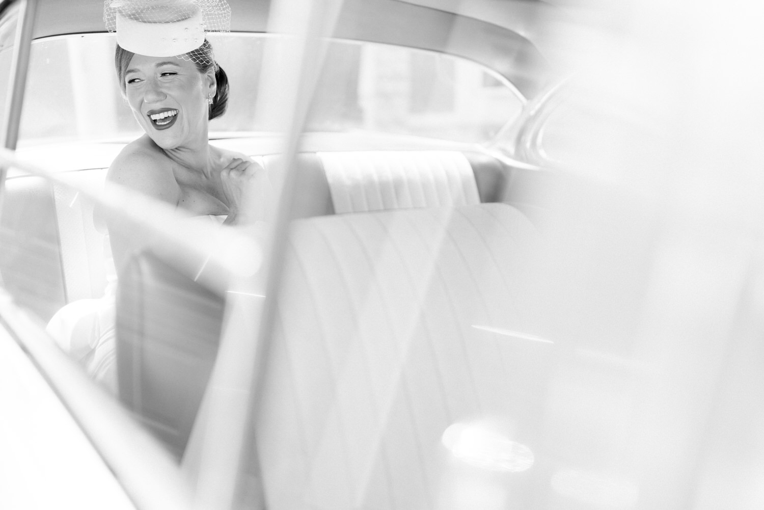 A smiling bride wearing a stylish veil, seated in the back of a vintage car, capturing a moment of happiness through the reflection of the window.