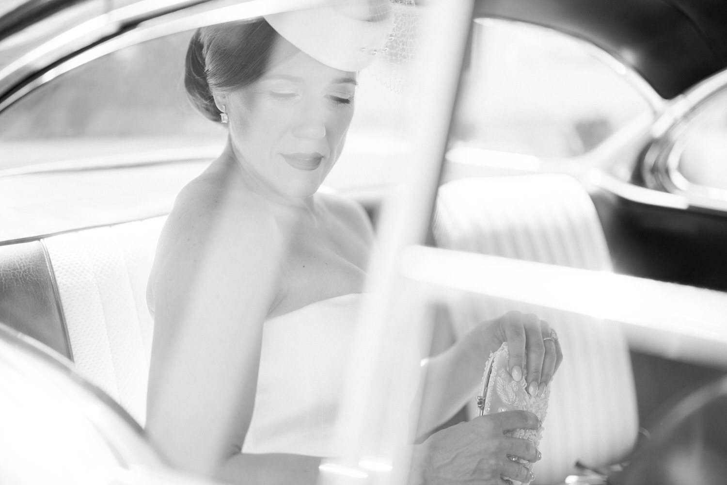 A serene bride in a strapless gown sits inside a vintage car, holding a beaded clutch, captured in soft black and white lighting.