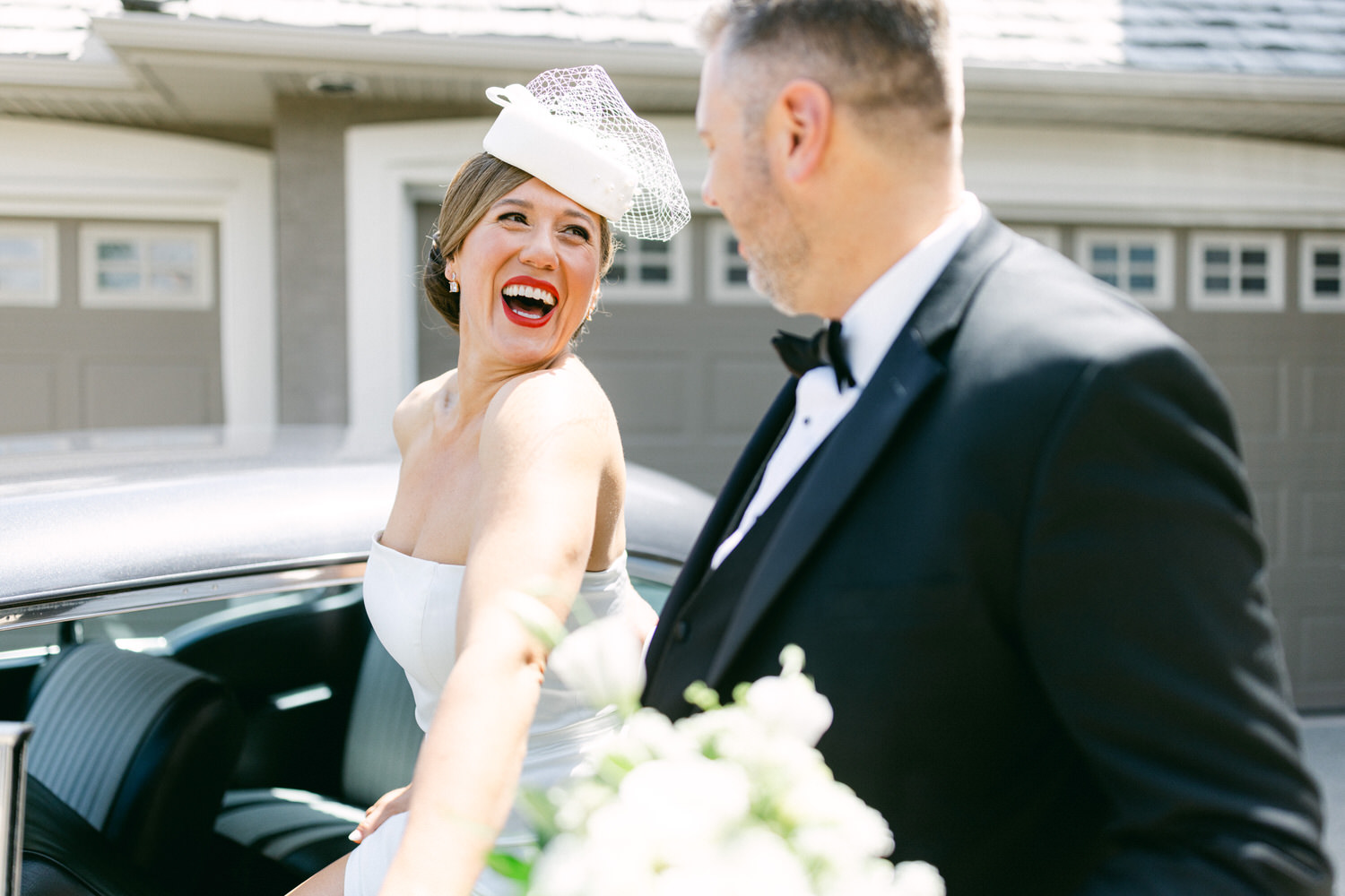 A bride in a white dress and hat shares a joyful moment with her partner outside a classic car, with a bouquet in her hand.