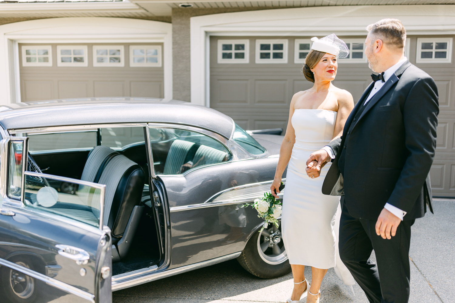 A bride in a sophisticated white dress and a groom in a tuxedo share a moment near a vintage car on a sunny day, showcasing their joy and style.