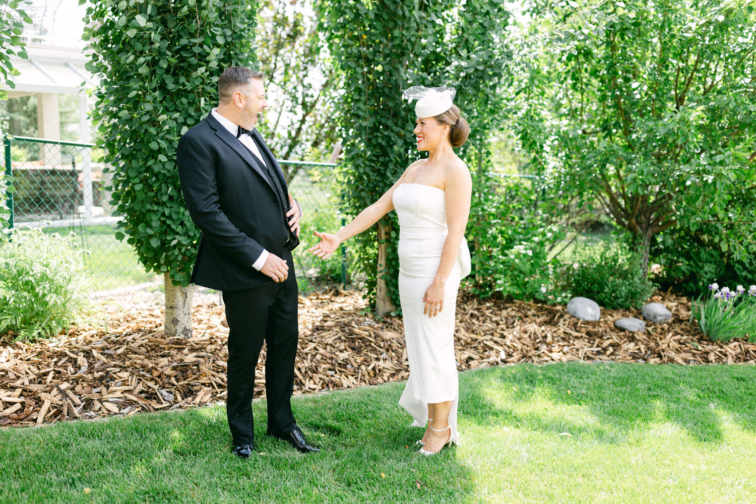 A man in a tuxedo and a woman in a stylish white dress engage in a joyful conversation in a vibrant garden setting.