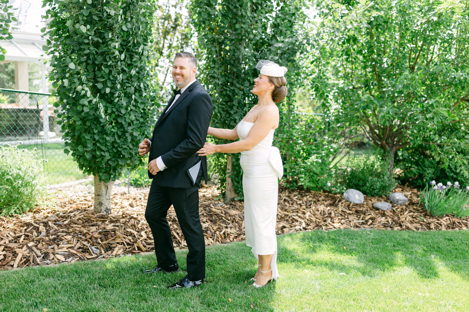 A bride playfully adjusting the groom's outfit outdoors, surrounded by lush greenery and natural light.