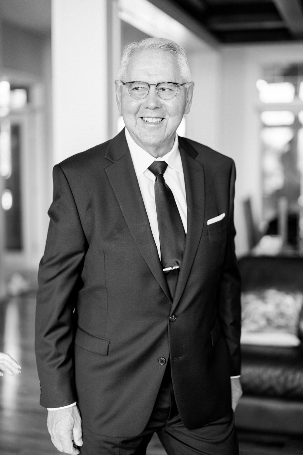 A gentleman in a black suit and tie, smiling warmly in a well-lit indoor setting.