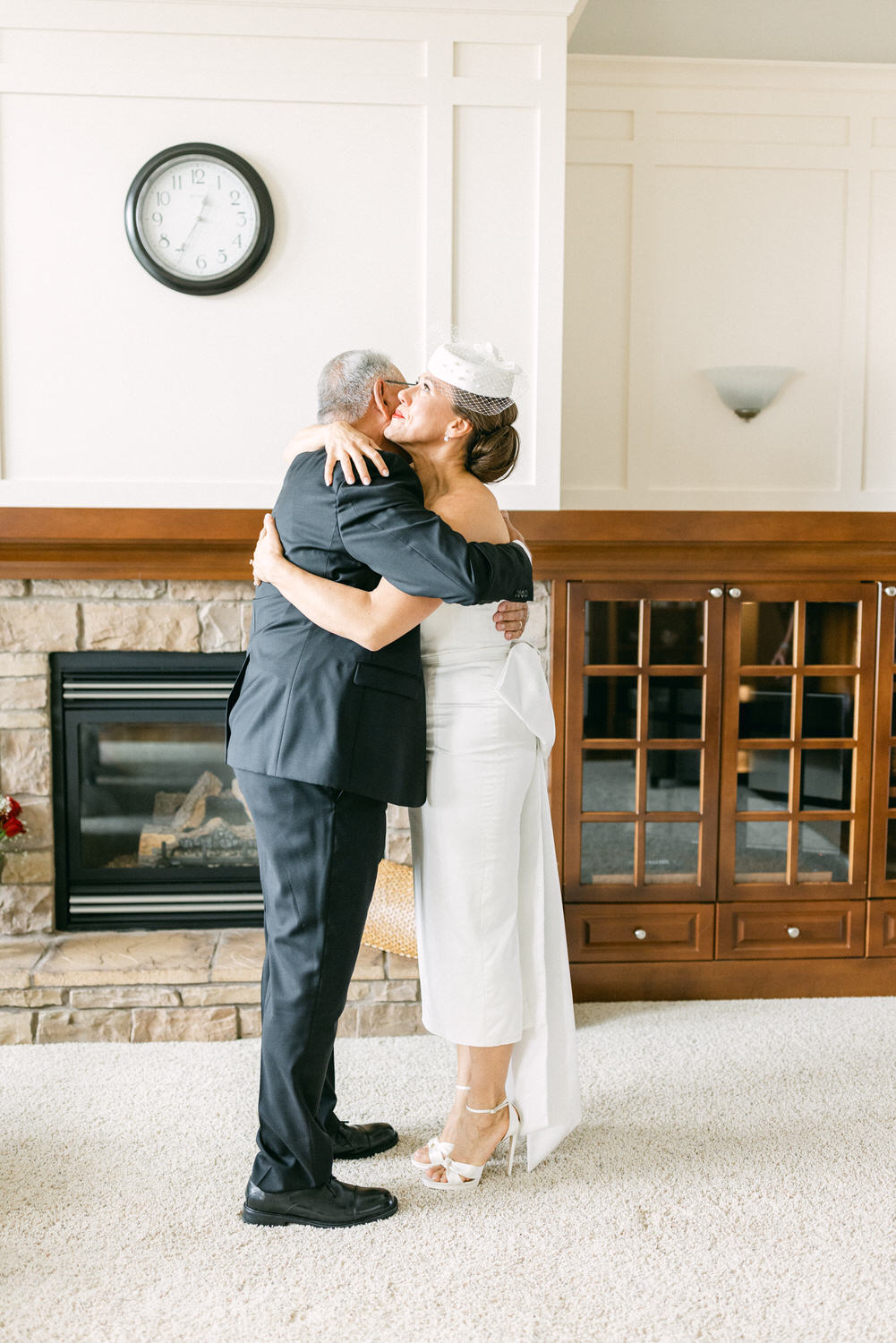 A bride and her father share a heartfelt hug in a cozy living room setting, with a clock on the wall and a fireplace in the background.