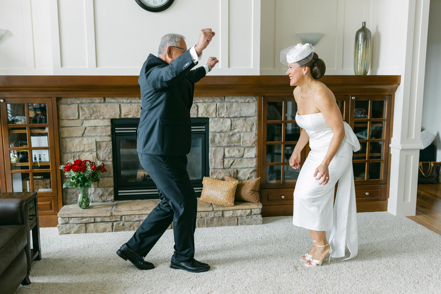 An elderly man and a woman in a white wedding dress dance playfully in a cozy living room, surrounded by cheerful decor and a beautiful flower arrangement.