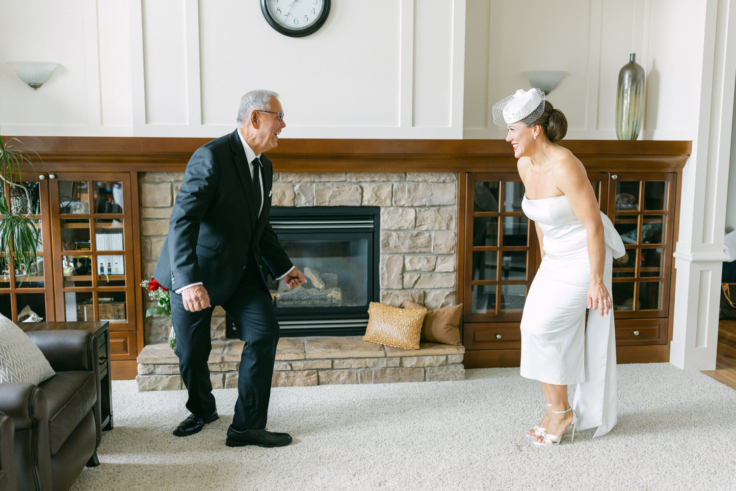 A smiling father in a suit dances joyfully with his daughter, who is wearing a white wedding dress, in a warmly decorated living room.