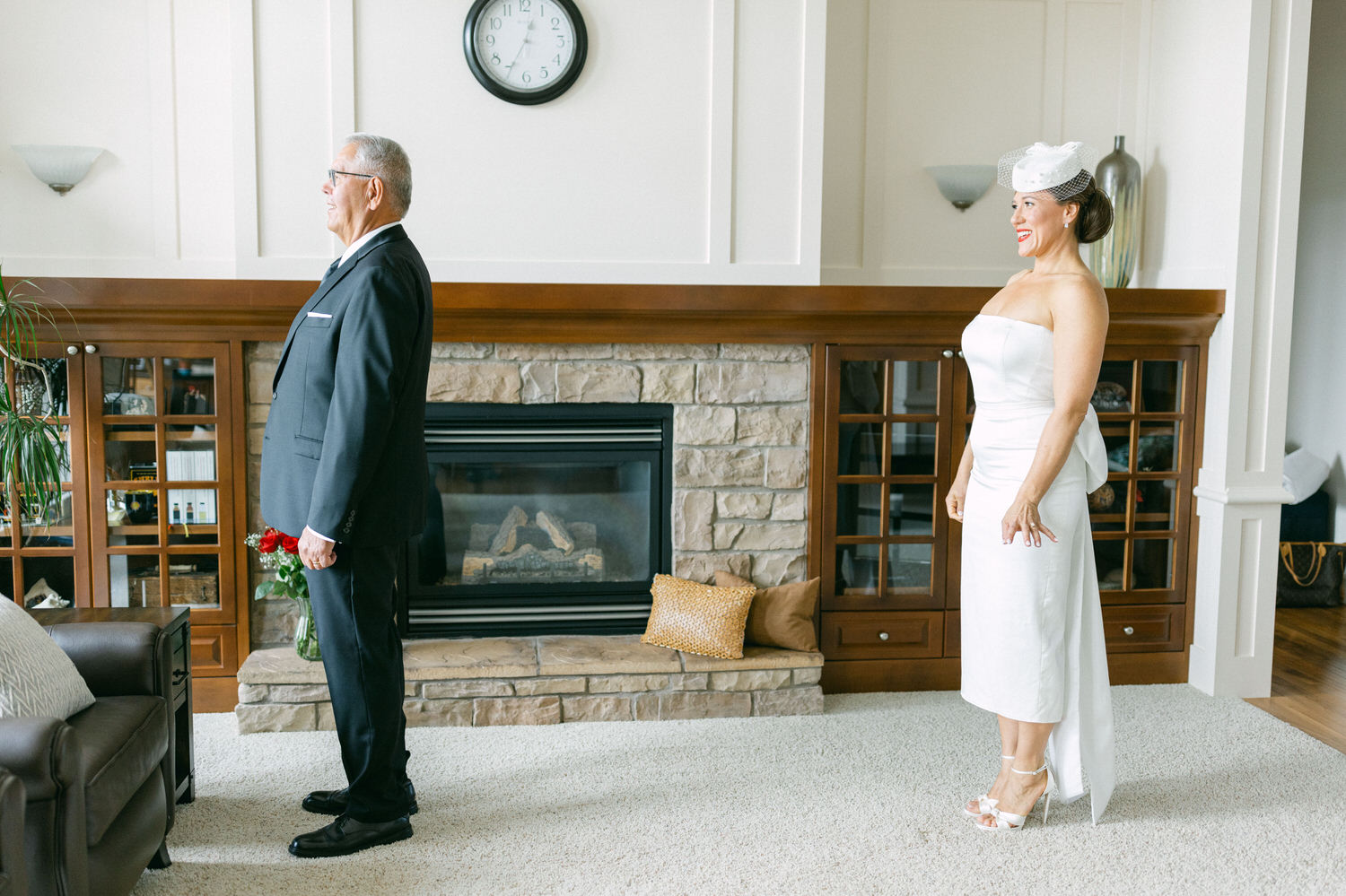 A bride in a white dress stands smiling, while a man in a black suit looks away, set against a cozy living room backdrop.