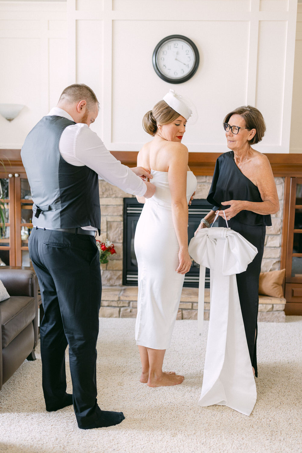 A bride in a white dress is being helped with her attire by a man and a woman in a cozy living room setting.