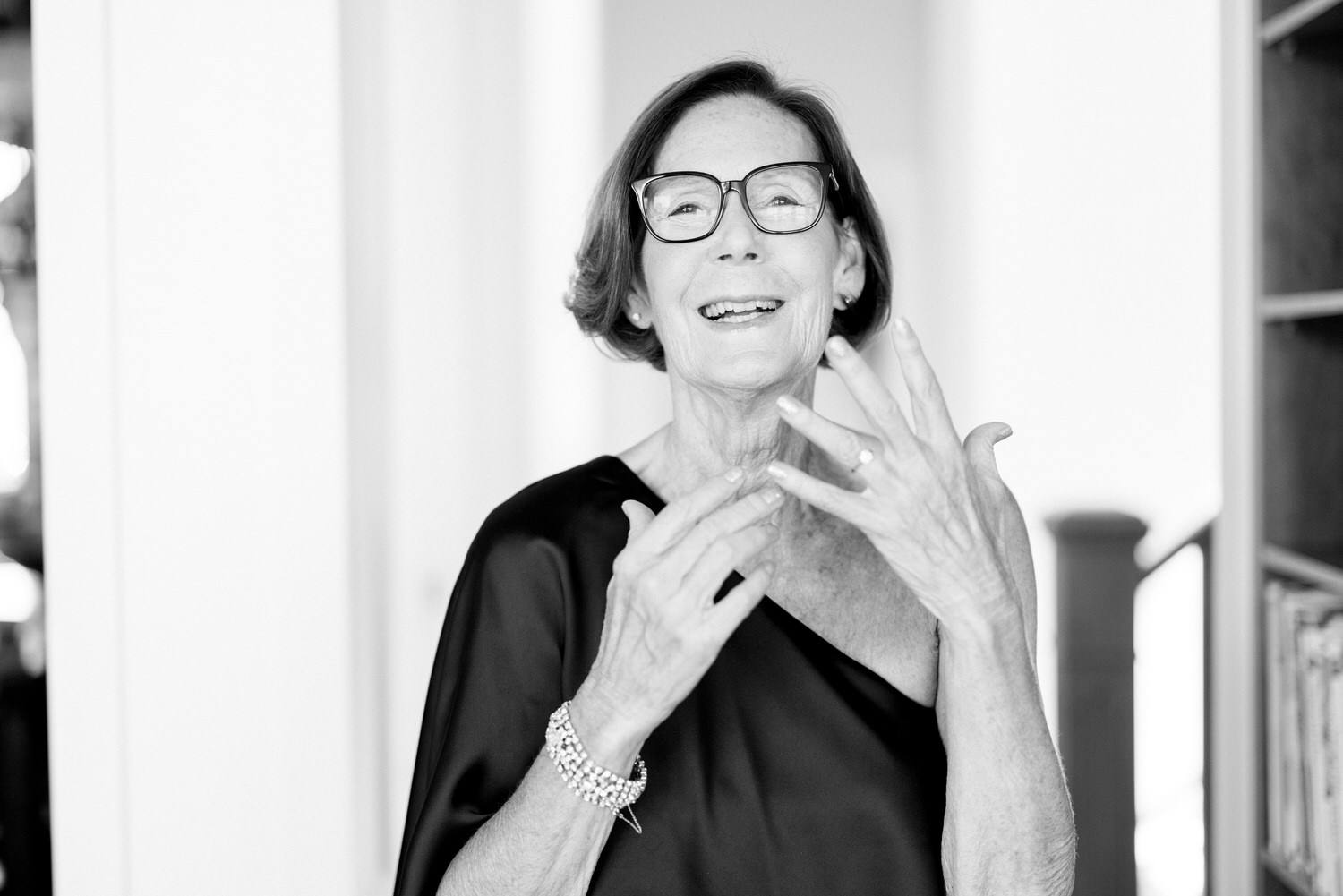 A smiling woman in a chic black dress joyfully gestures with her hands, showcasing her elegance against a softly lit backdrop.