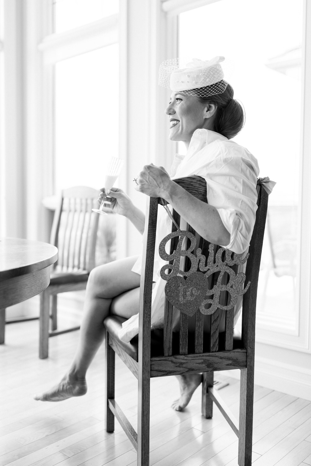 A joyful bride-to-be sitting in a wooden chair, holding a glass and wearing a white hat, with a decorative sign on the chair back.