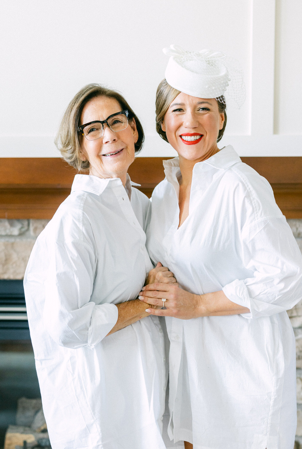 A smiling mother and daughter pose together in white shirts, showcasing their close bond in a bright, cozy setting. The daughter sports a unique white hat, adding elegance to the heartwarming scene.