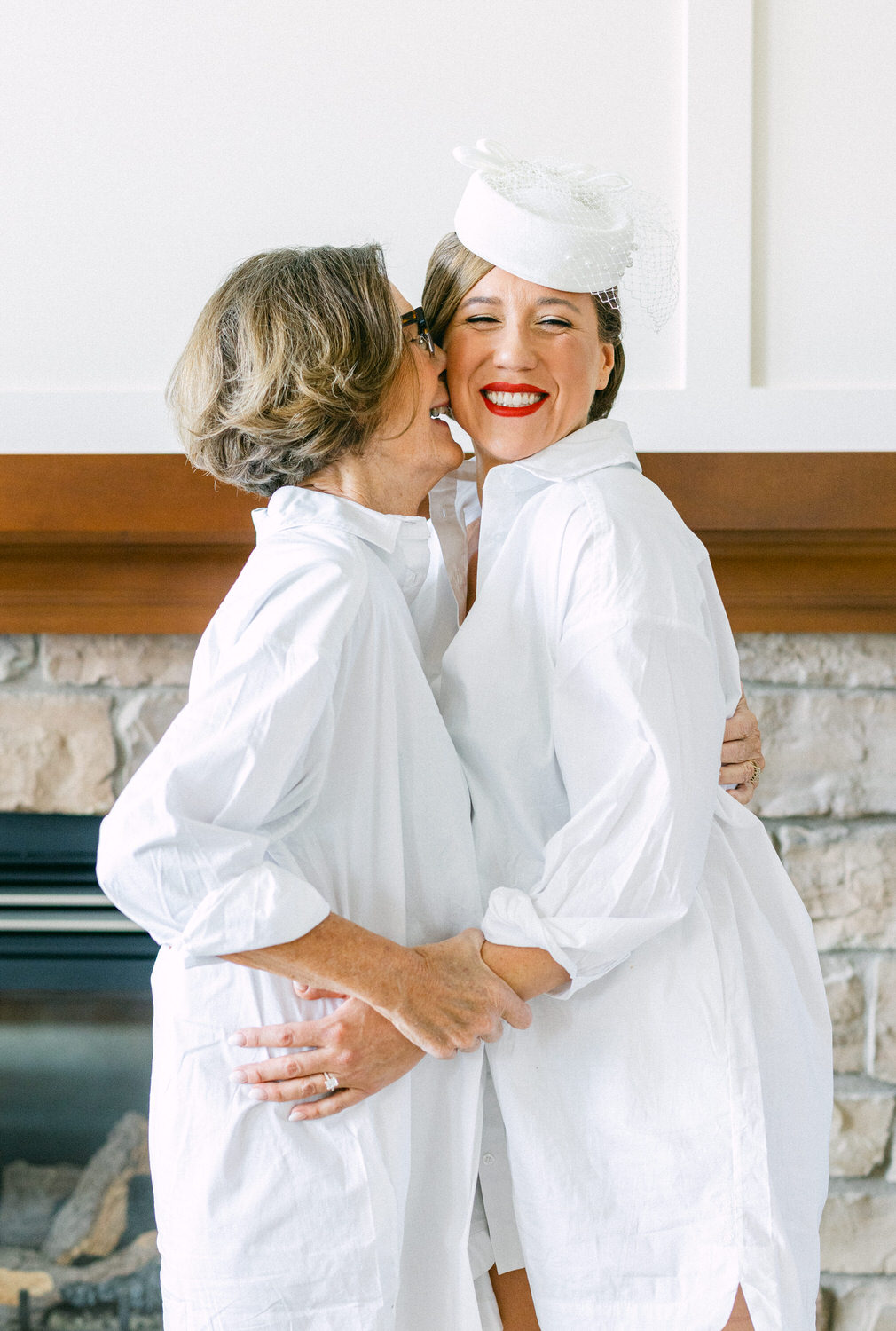 A joyful moment between two women in white shirts, celebrating together with laughter and warmth by a stone fireplace.