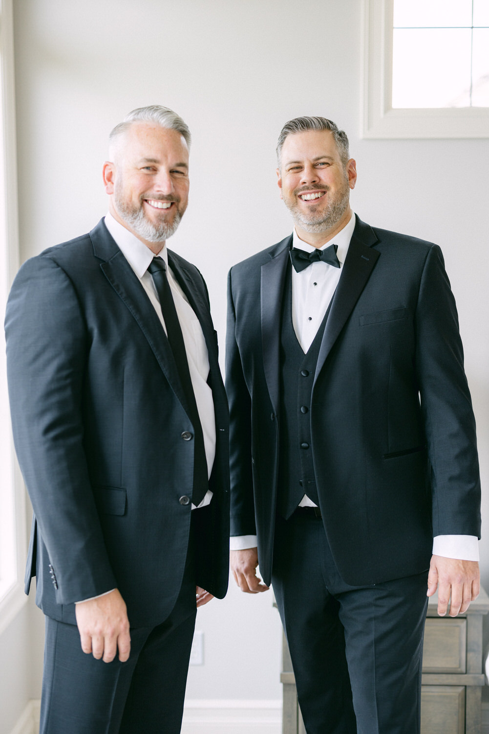 Two well-dressed men in black tuxedos smiling together in a well-lit interior setting.