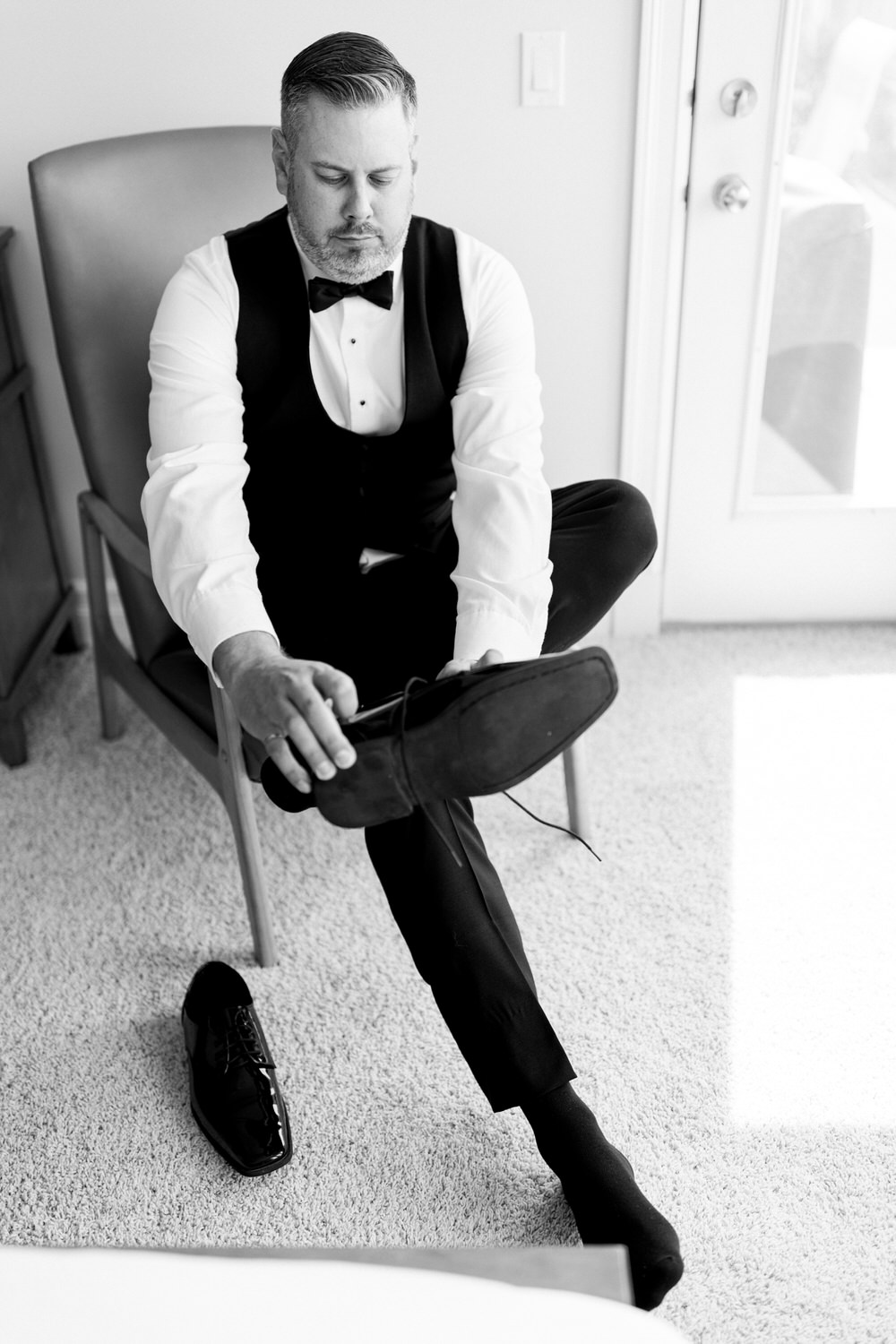 A man in formal attire sits on a chair while putting on a shoe, with a focused expression, surrounded by soft light and cozy interior details.