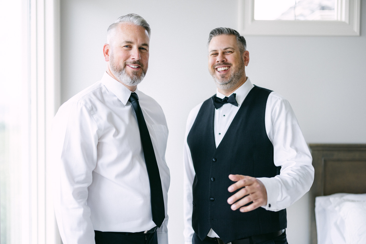 Two well-dressed men, one in a tuxedo and the other in a white shirt and black tie, smiling and posing in a bright room before a wedding.