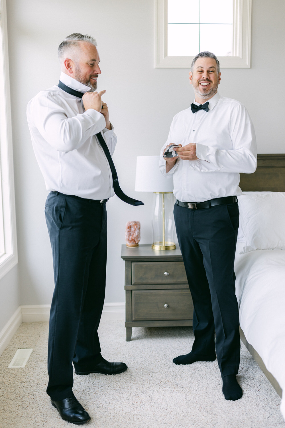 Two men adjusting their attire and looking cheerful while preparing for an event in a well-lit room.