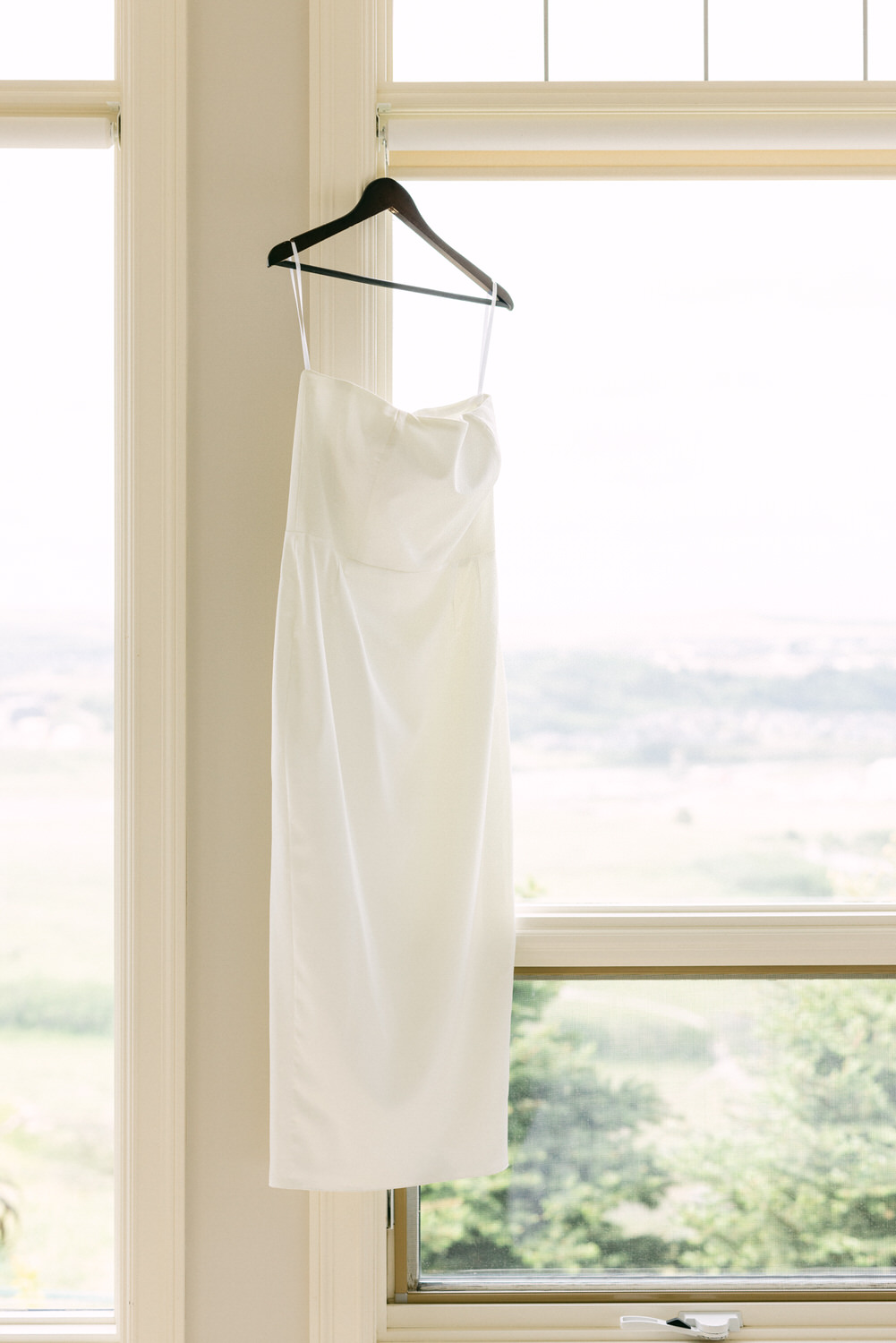 A simple white wedding dress draped elegantly on a hanger, illuminated by natural light streaming through a large window.