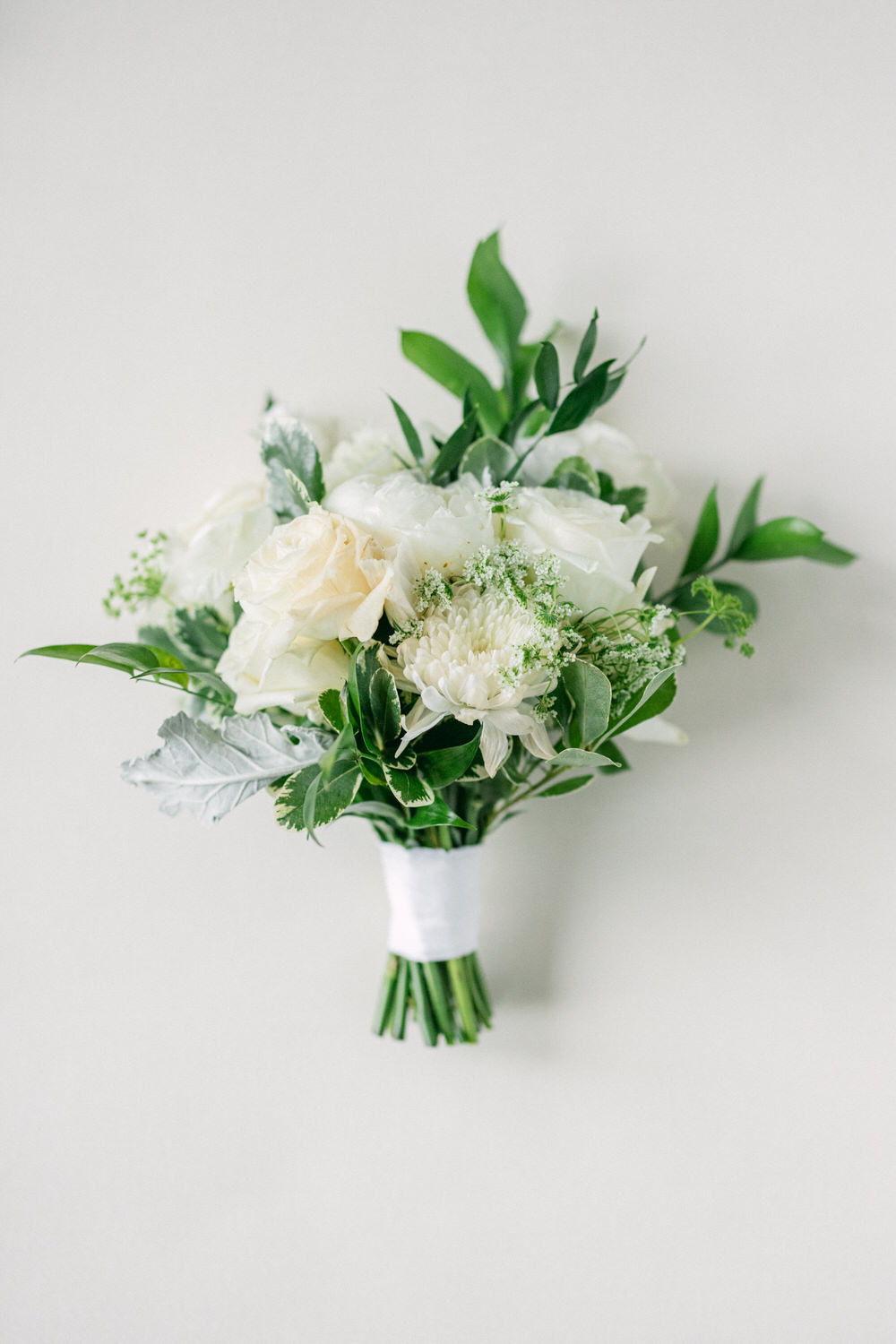 A beautifully arranged bouquet featuring white roses, greenery, and delicate flowers, wrapped in white ribbon against a neutral background.